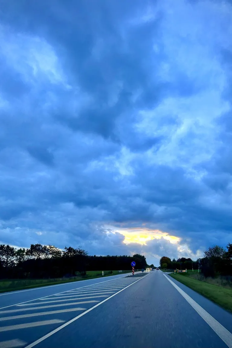 The road ahead as seen from the passenger seat in a car. Trees are lining the road ahead and in the distance a set of headlights are visible. In the horizon and above is a stormy sky with a tiny hole in the clouds.